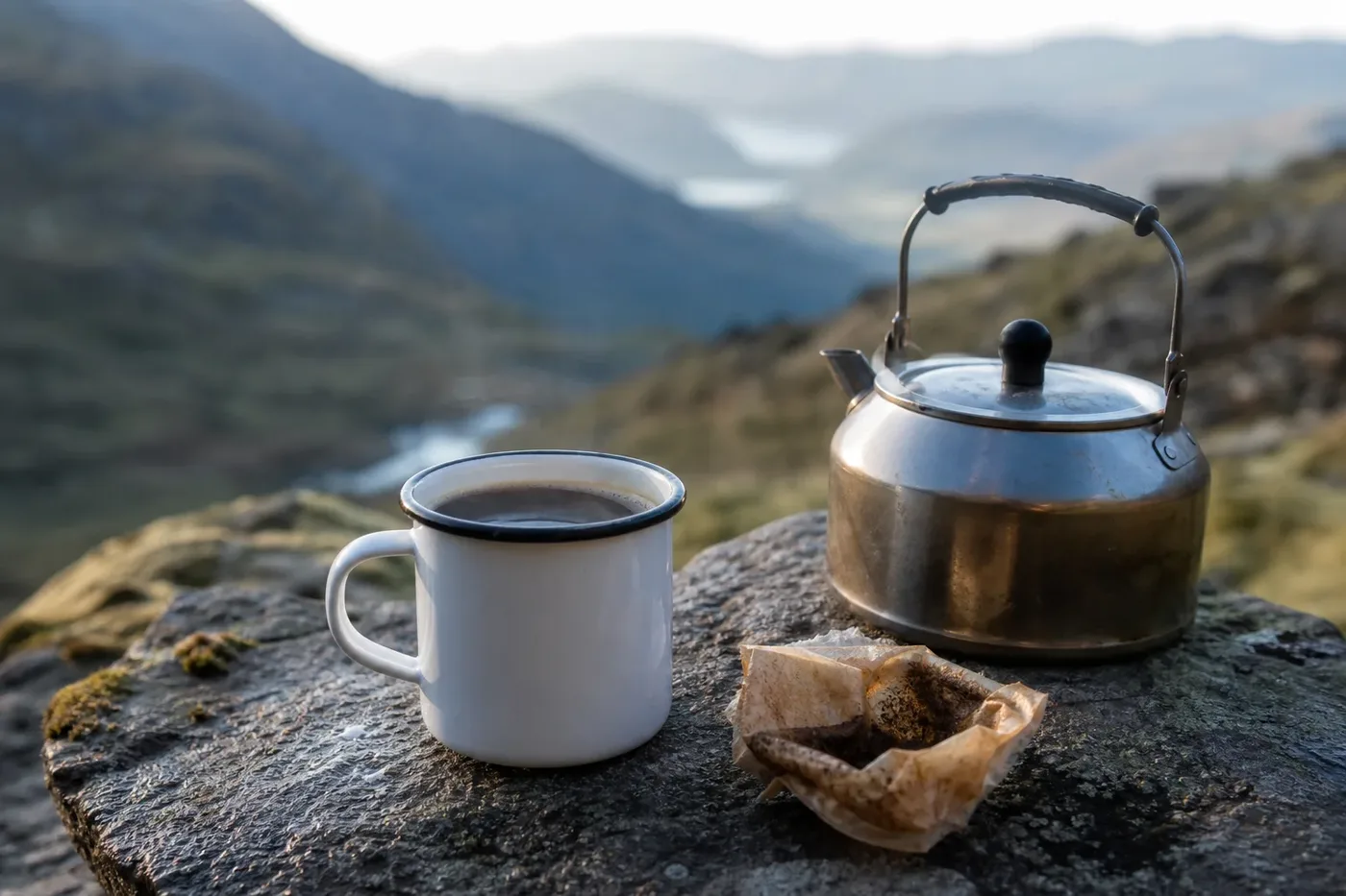 An enamel mug of coffee and a kettle on a rock outdoors, beside a torn-open coffee sachet