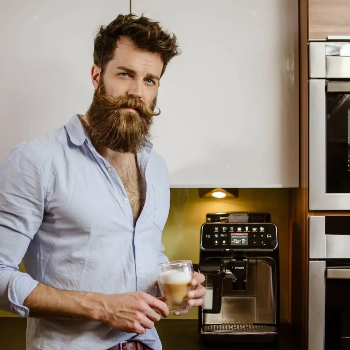 a person with a beard holding a glass of coffee in front of the Philips coffee machine