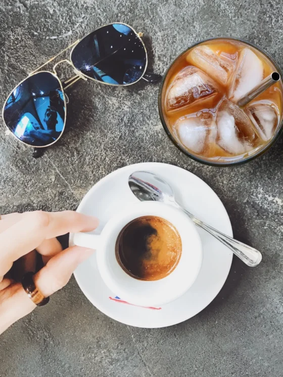 a hand holding a cup of coffee next to a glass of ice and sunglasses