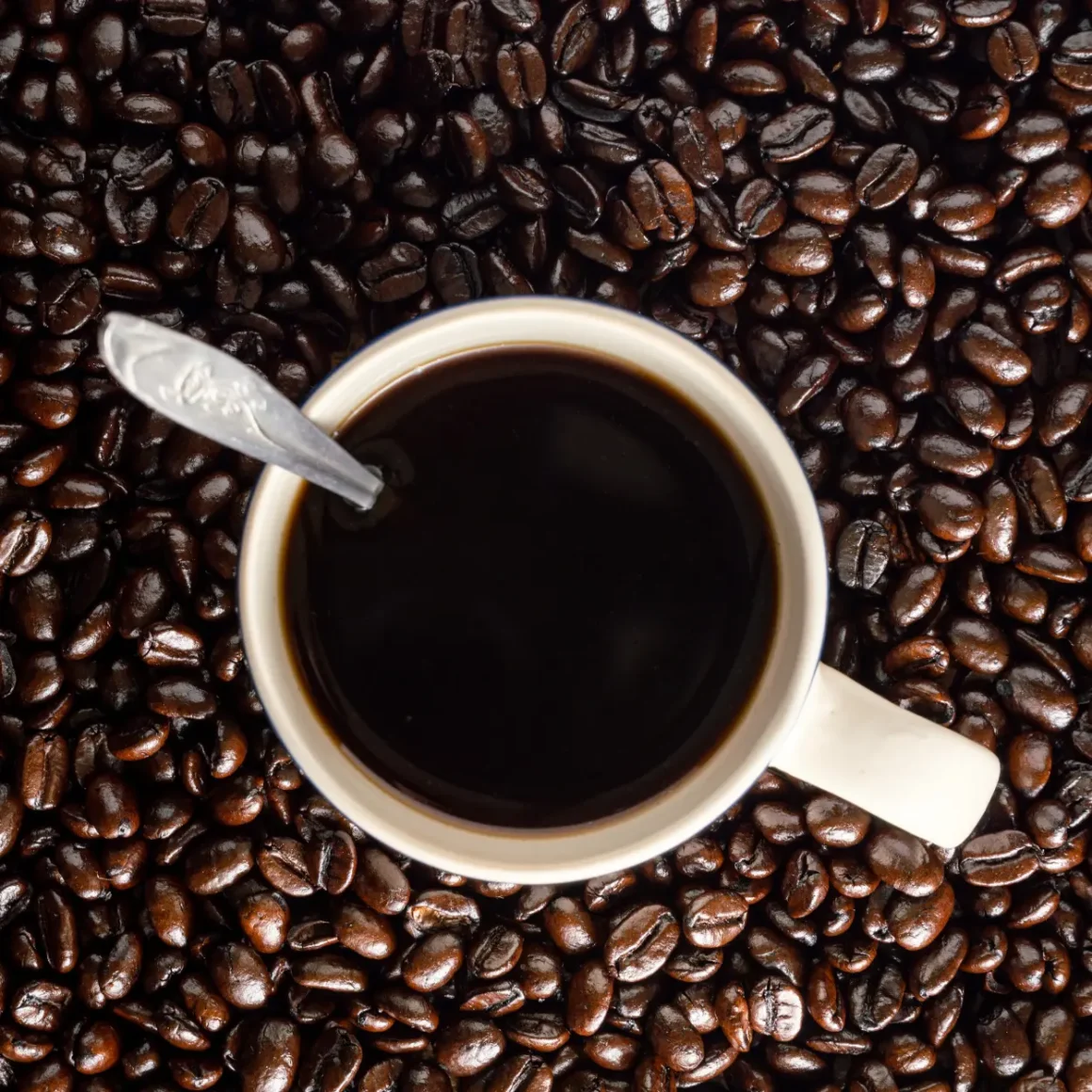 a cup coffee with a spoon over a pile of dark roast coffee beans