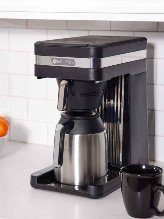a bunn 55200 CSB3T coffee maker with a black cup and a bowl of oranges on a counter