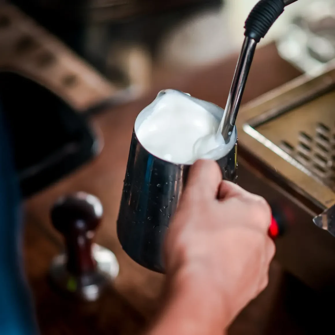 barista holding a metal cup and pouring milk from an espresso machine
