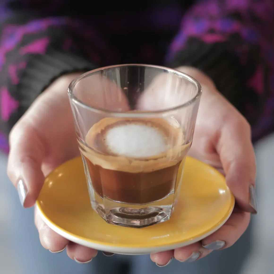 a person holding a small yellow plate with a glass of macchiato coffee