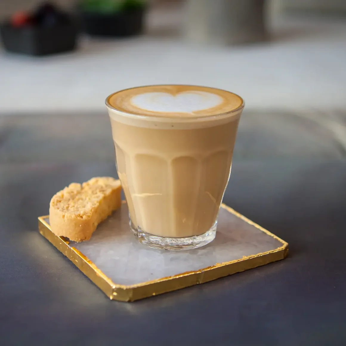 a glass of piccolo coffee with a heart design on top and a cookie on a tray