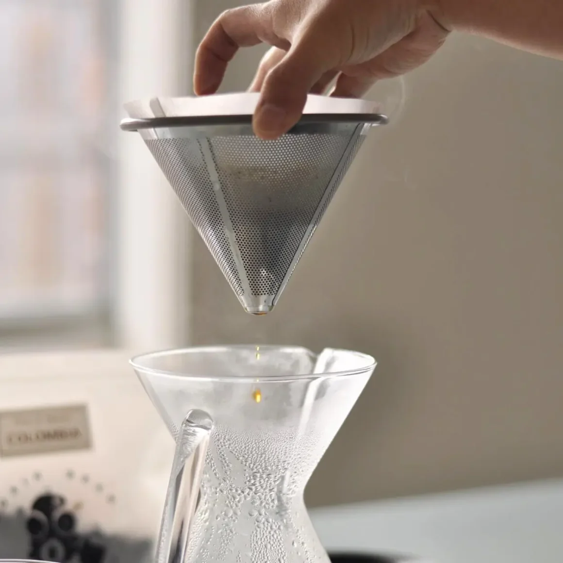a hand holding a ovalware paperless stainless steel chemex reusable pour over coffee filter over a glass container