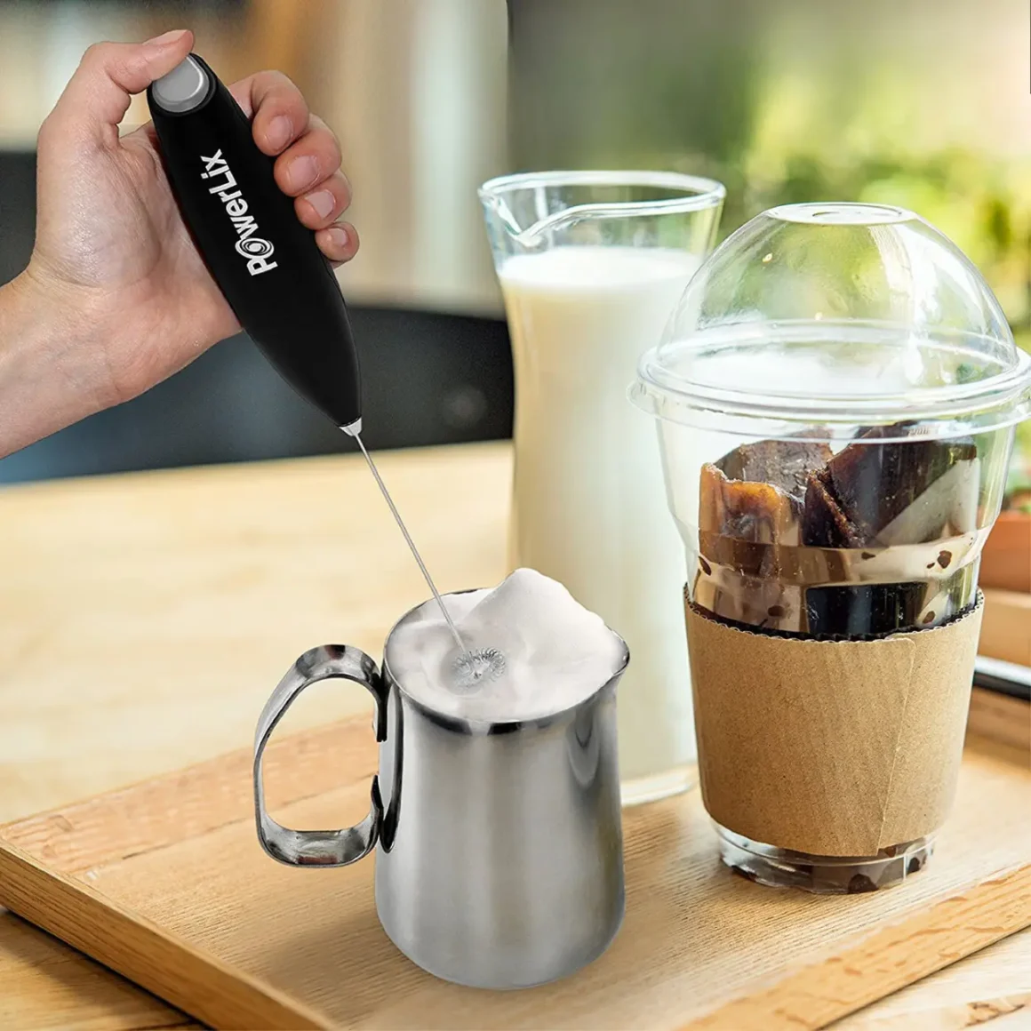 a hand holding a milk frother and a metal pitcher with a milk and a glass of milk