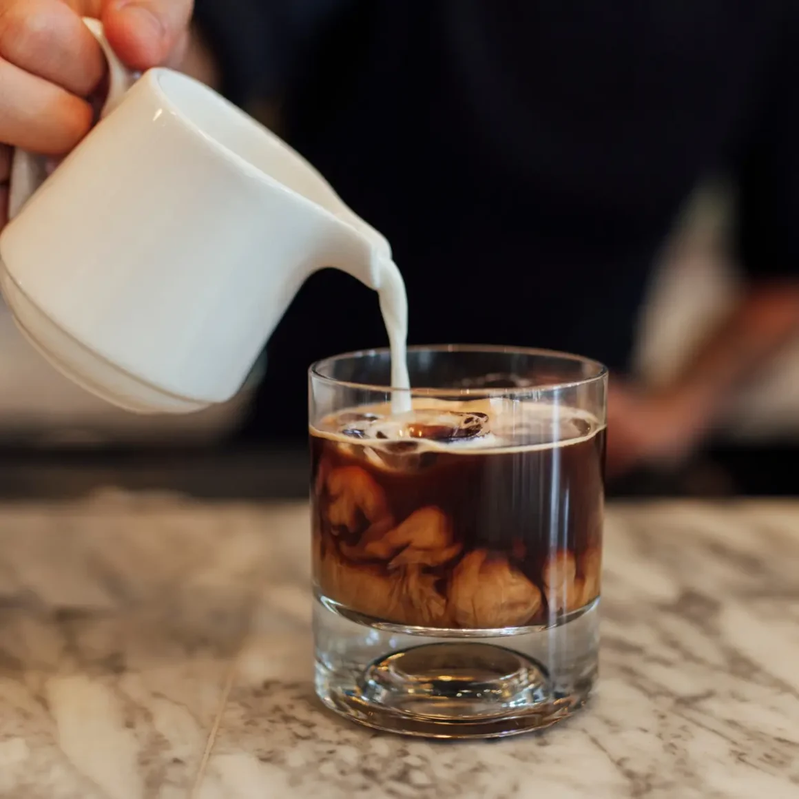 barista pouring milk into a glass of cold latte
